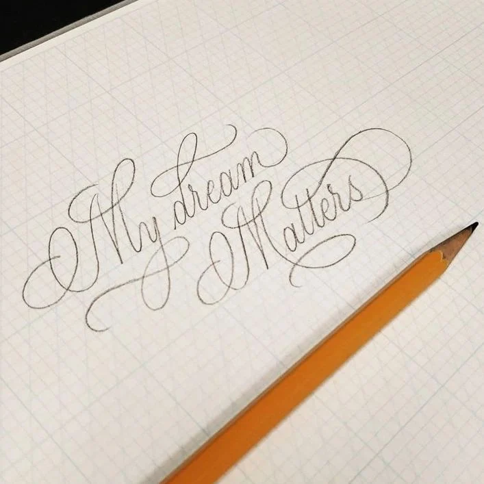 Pencil calligraphy spelling The Dream Makers with decorative flourishes on squared paper.