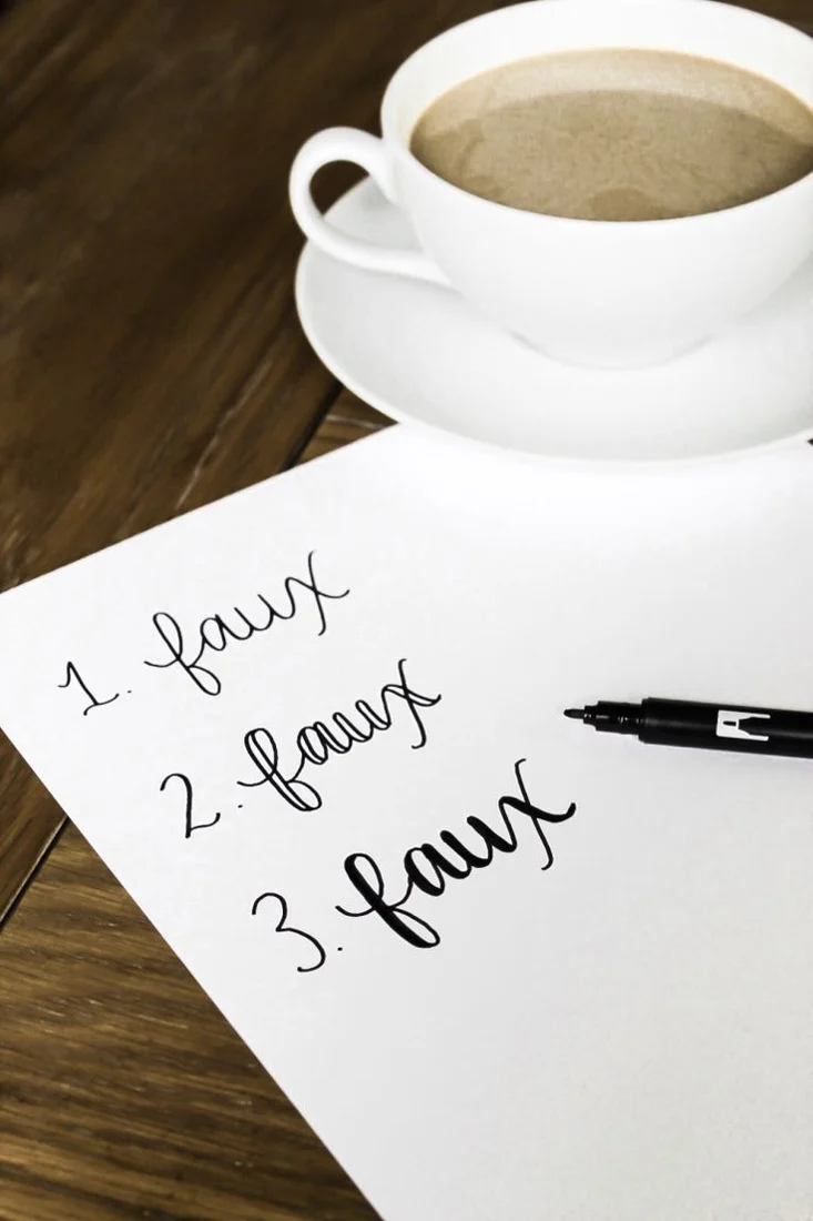 Three stages of faux calligraphy lettering on paper beside a black pen and a coffee cup.