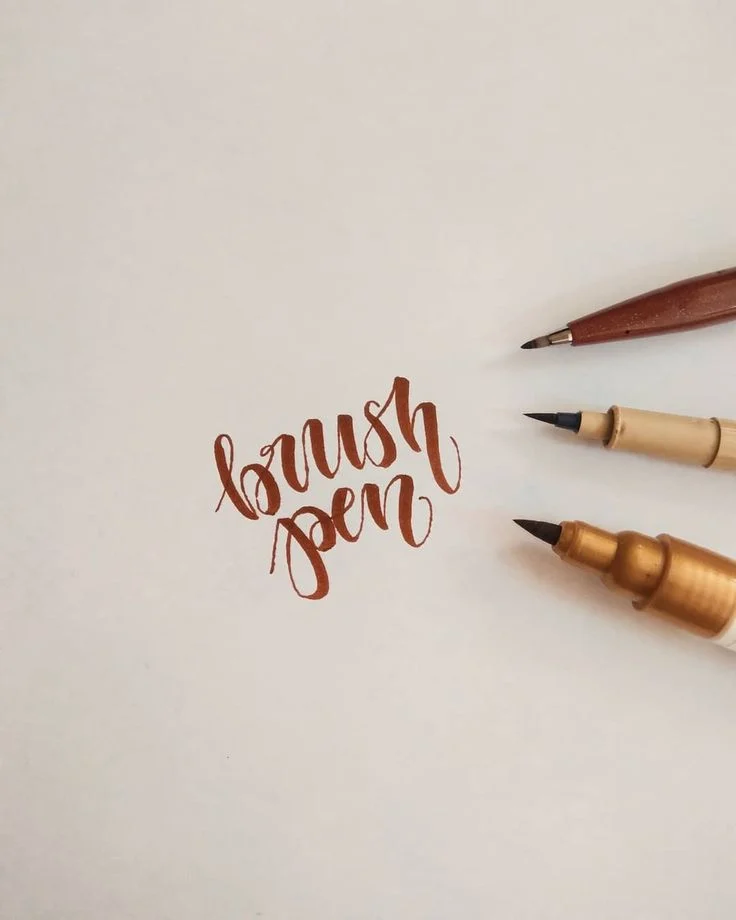 Brush pen calligraphy spelling brush pen in flowing brown lettering beside three pens.