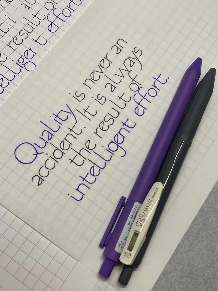 Monoline calligraphy practice in black and purple ink on grid paper beside two pens.