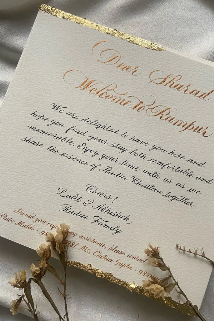 Hand-lettered welcome card in gold and black flowing script with gold edging and dried flowers.